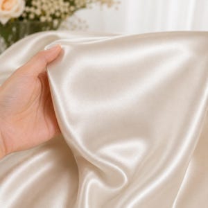 Op de afbeelding: Een stuk champagnekleurige stof met een gladde, glanzende en reflecterende satijnen afwerking. Het materiaal is gedrapeerd in zachte, vloeiende plooien, wat de glanzende textuur en de lichtreflecterende kwaliteit van het textiel benadrukt.