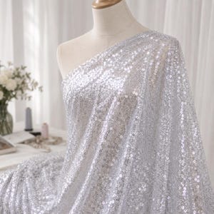 May include: A shimmering silver fabric covered in dense, reflective sequins draped over a mannequin. The material features a continuous, sparkling surface that catches the light, creating a bright, metallic appearance across the entire textile.