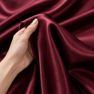 Puede incluir: Una tela de color burdeos profundo con una superficie lisa, brillante y reflectante. El material está drapeado en pliegues suaves y fluidos, mostrando un acabado de alto brillo que capta la luz, creando variaciones sutiles en el tono rojo.