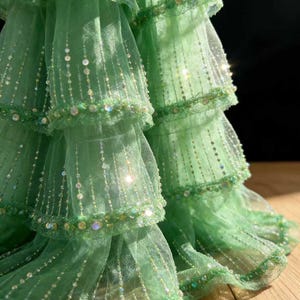 Op de afbeelding: Close-up van een gelaagde, lichtgroene stof met een delicate, transparante textuur. De stof is versierd met rijen glinsterende pailletten en kralen, wat een glinsterend effect creëert. De lagen zijn gerimpeld, wat volume en een vloeiende uitstraling geeft. Het ontwerp suggereert een formeel kledingstuk.