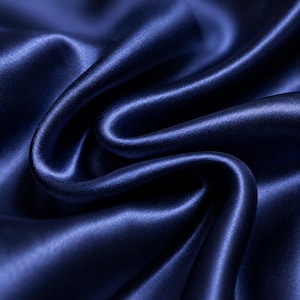 May include: A deep navy blue fabric with a smooth, lustrous finish and a reflective surface. The material is draped in soft, flowing folds that create subtle highlights and shadows, emphasising the dense, uniform texture of the dark blue textile.