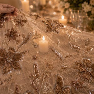 May include: A sheer, beige fabric with intricate floral embroidery and sequin embellishments. The design features large, detailed flowers and delicate leaf patterns. The fabric is draped, with a hand holding a portion of it, and lit by candlelight.