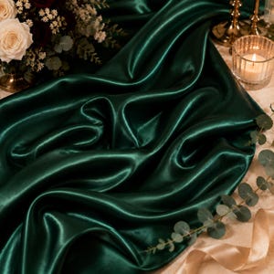 May include: A deep emerald green satin fabric runner draped in soft folds. The smooth, reflective material creates a rich texture with light highlights across the curves of the cloth, showcasing a lustrous finish and a vibrant, saturated color.