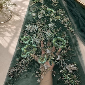 Puede incluir: Una tela de tul verde oscuro con intrincados bordados florales con lentejuelas y cuentas en verde claro, verde oscuro y oro metálico. El diseño muestra patrones botánicos detallados con pétalos en capas y tallos en forma de enredadera.