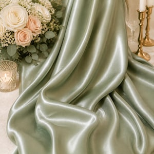 May include: A sage green satin fabric draped in soft folds, accompanied by a bouquet of white and pale pink roses, a glass candle holder with a lit candle, and two gold-toned metal candlesticks with white taper candles.