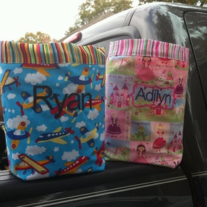 May include: Two fabric tote bags. One is blue with airplanes and the name "Ryan" embroidered in black. The other is pink with princess-themed squares and the name "Adilyn" embroidered in blue. Both have striped trim.