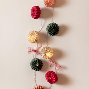 Könnte beinhalten: Eine Lichterkette mit dekorativen Papierkugeln in verschiedenen Farben, darunter Rot, Grün und Gold. Die Lichter sind auf einem dünnen Silberdraht aufgereiht und mit kleinen rosa Bändern verziert. Ideal für Feiertagsdekorationen.