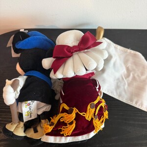 May include: Two plush Disney character dolls. One doll is dressed in a black and blue outfit with a walking stick. The other doll is wearing a white hat with a red bow and a red velvet dress with gold tassels. A white flag is visible.