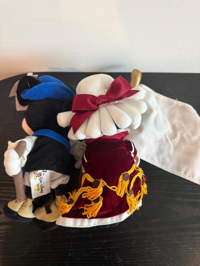 May include: Two plush Disney character dolls. One doll is dressed in a black and blue outfit with a walking stick. The other doll is wearing a white hat with a red bow and a red velvet dress with gold tassels. A white flag is visible.