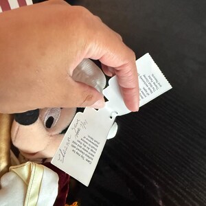 May include: A plush Mickey Mouse doll in a gold and burgundy uniform with a white collar. The doll has a tag with handwritten text and care instructions. The doll's face is visible, with black eyes and a white muzzle.