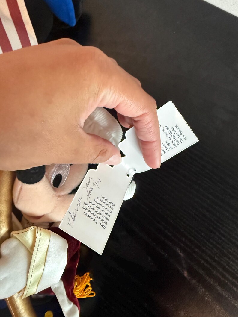 May include: A plush Mickey Mouse doll in a gold and burgundy uniform with a white collar. The doll has a tag with handwritten text and care instructions. The doll's face is visible, with black eyes and a white muzzle.