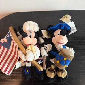 May include: Two plush Mickey Mouse figures in patriotic outfits. One holds a flag with red and white stripes and a gold staff. The other has a blue hat and a drum. Both have black shoes with flower accents.