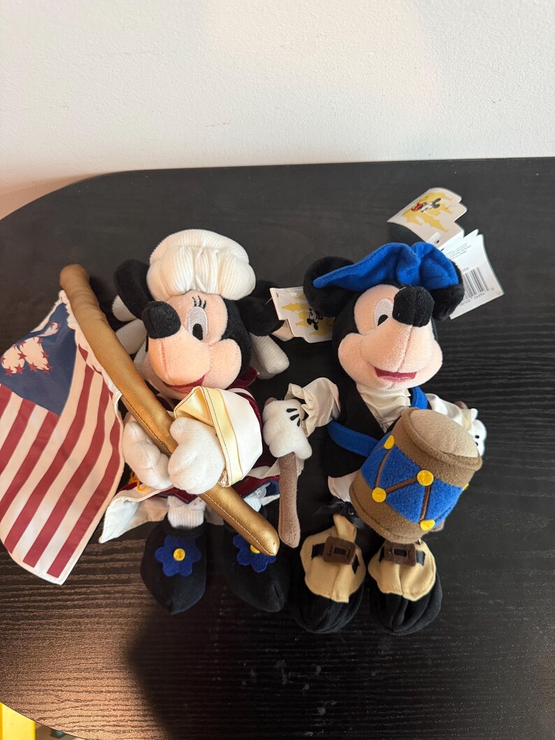May include: Two plush Mickey Mouse figures in patriotic outfits. One holds a flag with red and white stripes and a gold staff. The other has a blue hat and a drum. Both have black shoes with flower accents.