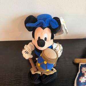 May include: A Mickey Mouse soft toy wearing a blue colonial hat and vest, holding a drum. The toy has black ears, a white face, and black shoes. A tag is attached to the toy.