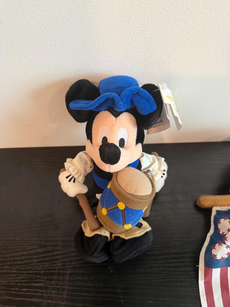 May include: A Mickey Mouse soft toy wearing a blue colonial hat and vest, holding a drum. The toy has black ears, a white face, and black shoes. A tag is attached to the toy.