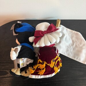 May include: Two plush figures of Mickey and Minnie Mouse in formal outfits. Mickey wears a blue hat, black jacket, and white trousers. Minnie is in a white hat with a red bow and a red velvet dress with gold tassels. A white flag is visible.