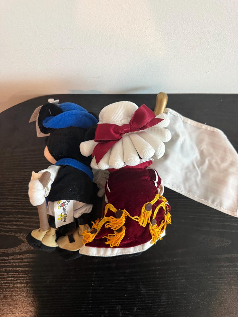 May include: Two plush figures of Mickey and Minnie Mouse in formal outfits. Mickey wears a blue hat, black jacket, and white trousers. Minnie is in a white hat with a red bow and a red velvet dress with gold tassels. A white flag is visible.