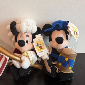 May include: Two plush Mickey Mouse and Minnie Mouse dolls. Minnie Mouse is dressed in a colonial outfit with a white hat and a red and gold outfit. Mickey Mouse is dressed in a blue hat and a blue and black outfit, holding a drum.