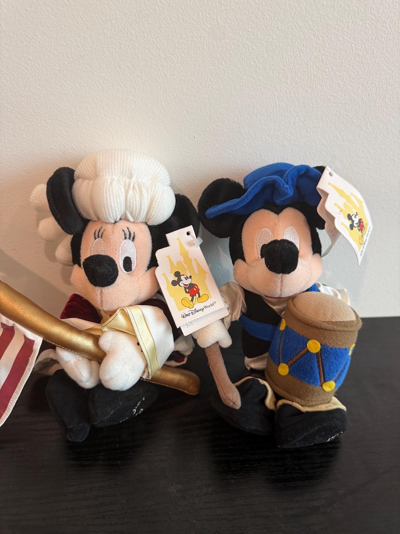 May include: Two plush Mickey Mouse and Minnie Mouse dolls. Minnie Mouse is dressed in a colonial outfit with a white hat and a red and gold outfit. Mickey Mouse is dressed in a blue hat and a blue and black outfit, holding a drum.