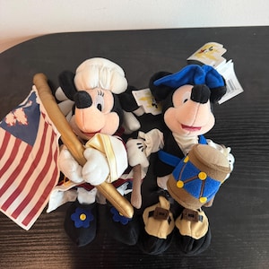 May include: Two plush Mickey Mouse figures in patriotic outfits. One holds a flag, the other a drum. Both have black ears, red shorts, and white gloves. One wears a white hat, the other a blue hat.