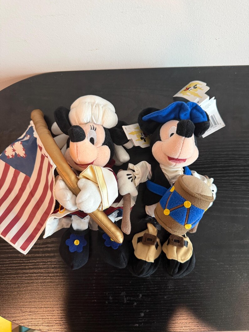 May include: Two plush Mickey Mouse figures in patriotic outfits. One holds a flag, the other a drum. Both have black ears, red shorts, and white gloves. One wears a white hat, the other a blue hat.