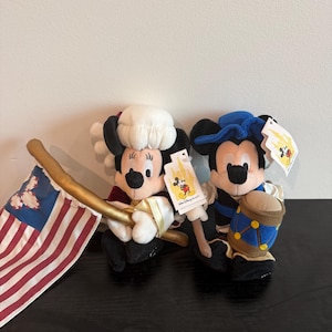 May include: Two plush Mickey Mouse and Minnie Mouse figures in patriotic outfits. Minnie Mouse holds a flag with red and white stripes and blue stars. Mickey Mouse holds a drum. Both figures have tags attached.