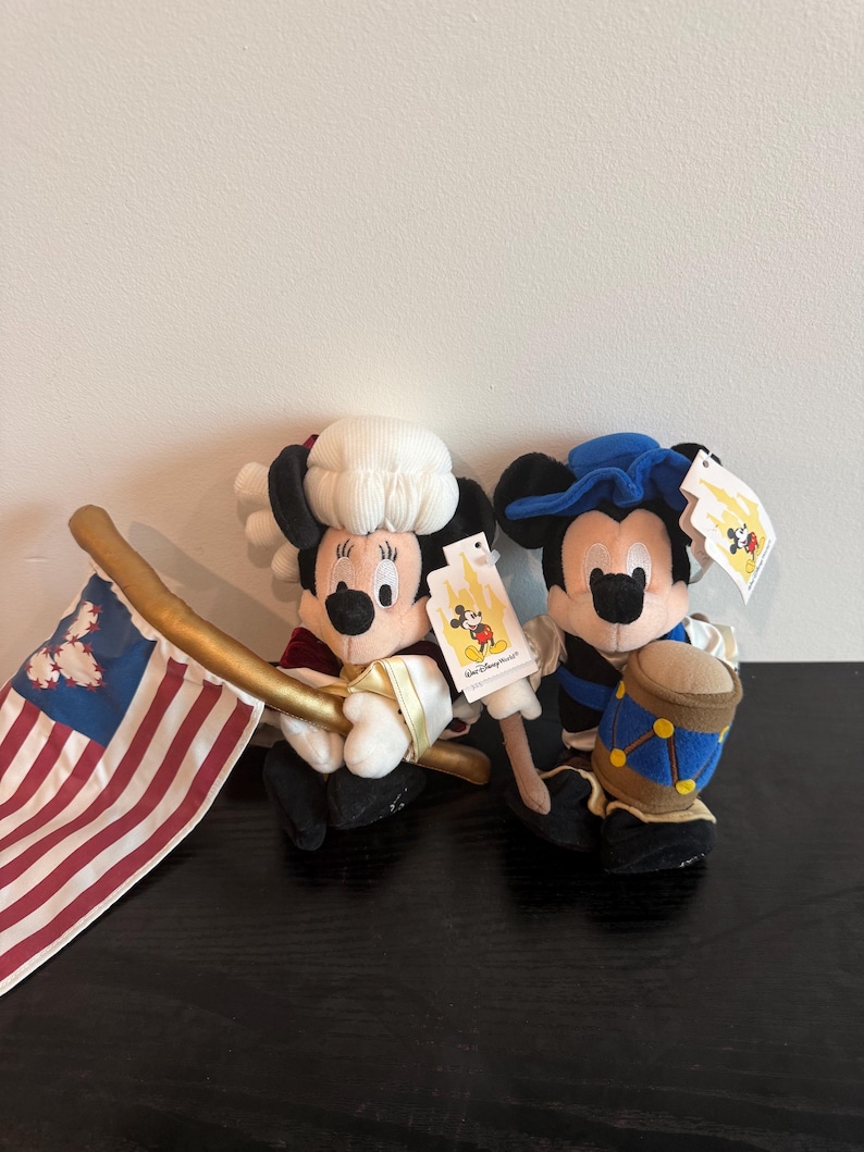 May include: Two plush Mickey Mouse and Minnie Mouse figures in patriotic outfits. Minnie Mouse holds a flag with red and white stripes and blue stars. Mickey Mouse holds a drum. Both figures have tags attached.