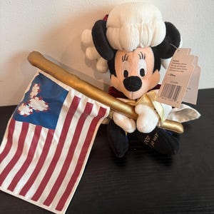 May include: A plush Minnie Mouse doll in colonial dress, holding a gold staff and a small American flag. The flag has red and white stripes with a blue corner. The doll wears a white wig and a red and white outfit. The doll is a patriotic item.