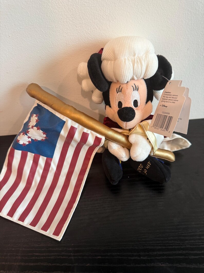 May include: A plush Minnie Mouse doll in colonial dress, holding a gold staff and a small American flag. The flag has red and white stripes with a blue corner. The doll wears a white wig and a red and white outfit. The doll is a patriotic item.