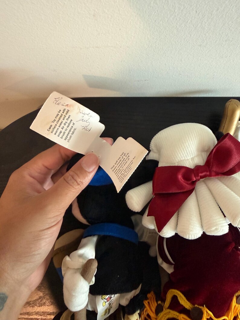 May include: Two plush dolls, one in a black outfit with a blue collar and white gloves, and the other in a red and white outfit. Both dolls have tags attached. The tag on the left reads "Care: Toy may be surface cleaned with warm water and mild soap."