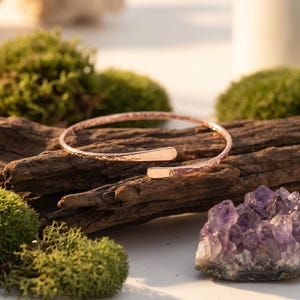May include: A rose gold open cuff bracelet with a hammered texture. The bracelet is displayed on a piece of driftwood, with green moss and a purple amethyst crystal in the background. The bracelet's design is simple and elegant.