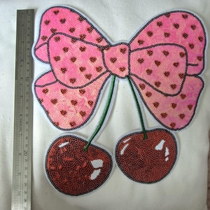 May include: A sequined patch featuring a large pink bow with brown heart patterns attached to two red cherries with green stems. A metal ruler placed to the left shows the patch measures approximately 23 cm in total height.