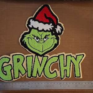 May include: A tufted textile patch featuring a green character wearing a red and white Santa hat above the text GRINCHY in green letters with black outlines and gold borders. A metal ruler shows the design is approximately 30 cm wide.