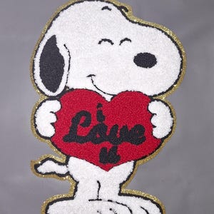 May include: A Snoopy character patch holding a red heart with the word "Love" in black. The patch has a white, textured body, black ears and spots, and a gold glitter border. The background is a solid grey.