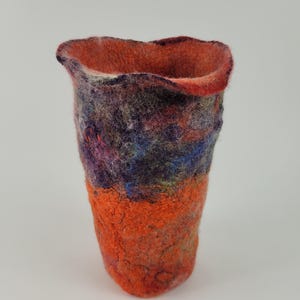 Könnte beinhalten: Ein Gefäß aus gefilzter Wolle mit einer strukturierten, unebenen Oberfläche. Die untere Hälfte ist leuchtend orange, während die obere Hälfte eine Mischung aus tiefvioletten, blauen und dunkelroten Fasern aufweist. Der Rand ist wellig und unregelmäßig.