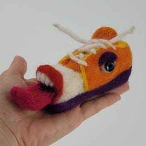 Könnte beinhalten: Ein skurriler Filz-Sneaker in Orange, Weiß und Lila, mit einer herausragenden rosa Zunge und einem blauen Auge. Der Schuh hat weiße Schnürsenkel und gelbe Akzente, was ein verspieltes, künstlerisches Design ergibt.