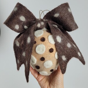 Könnte beinhalten: Handgefertigter Weihnachtsbaumschmuck aus Filz. Der Schmuck ist eine Kugel mit einer braunen Schleife, verziert mit weißen Punkten. Die Kugel selbst ist beige mit weißen und braunen Punkten. Eine Schnur ist zum Aufhängen befestigt.