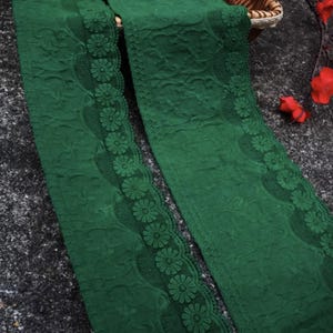 Puede incluir: Dos tiras de tela verde oscuro con un ribete de encaje floral. La tela tiene un patrón texturizado y el ribete de encaje presenta un diseño de margarita repetido. Las tiras están dispuestas diagonalmente sobre un fondo oscuro.