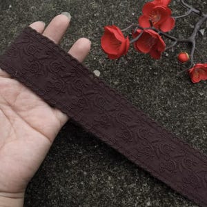 Puede incluir: Cinta marrón oscuro con un patrón floral y bordes festoneados. La cinta se sostiene en una mano, con flores rojas en el fondo. La cinta mide aproximadamente 3,8 cm de ancho.