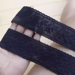 Puede incluir: Dos tiras de encaje negro con un patrón floral. El encaje tiene un borde festoneado y se sostiene en una mano. El encaje se utiliza probablemente para coser, hacer manualidades o añadir detalles a la ropa.