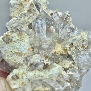 Puede incluir: Primer plano de un grupo de cristales de cuarzo transparente. Los cristales puntiagudos están incrustados en una matriz de roca beige y verde claro. El cuarzo tiene un aspecto vítreo. Formación natural con estética terrosa.