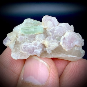 Puede incluir: Un grupo de gemas en bruto con un cristal de turmalina verde pálido y varios cristales de turmalina rosa. Los cristales están engastados en una matriz de cuarzo blanco y translúcido. El fondo es oscuro.