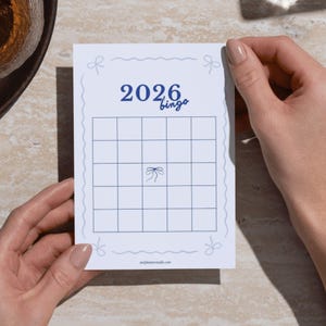 Peut inclure: Une carte de bingo blanche avec une grille bleue et le texte "2026 bingo". La carte est ornée d'accents bleus en forme de nœuds et tenue par deux mains. L'adresse du site web est imprimée en bas de la carte.