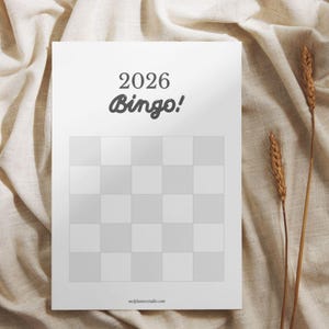 Peut inclure: Une carte de Bingo blanche avec le texte "2026 Bingo!" dans une police ludique. La carte présente une grille de cases, certaines ombragées en gris clair. Deux épis de blé sont placés sur le côté droit de la carte, sur un fond de tissu beige.