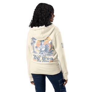 Può includere: Felpa con cappuccio color crema con una grafica sul retro raffigurante un gallo in uniforme da Jiu Jitsu, palme e le parole "De Boa Jiu Jitsu". La felpa ha un cappuccio con coulisse e una tasca a marsupio.