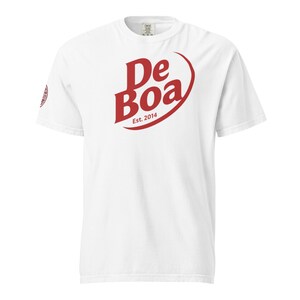 Może przedstawiać: Biała koszulka z czerwonym logo "De Boa" i "Est. 2014". Logo jest w odważnej, stylizowanej czcionce z zakrzywionym elementem projektu. Koszulka ma krótkie rękawy i okrągły dekolt.