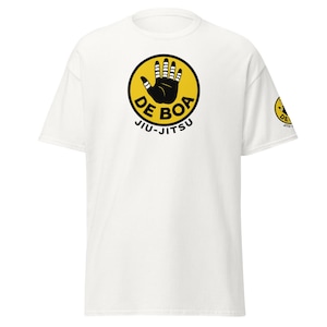 Może przedstawiać: Biała koszulka z żółtym, okrągłym logo przedstawiającym czarną dłoń i napisem "DE BOA JIU-JITSU". Mniejsza wersja logo znajduje się na rękawie. Koszulka wykonana jest z miękkiego materiału.
