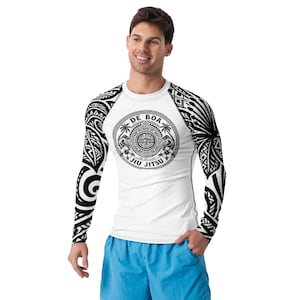 May include: White long-sleeve rash guard with black tribal arm designs and a circular graphic on the chest. The graphic reads "DE BOA JIU JITSU" with palm trees and floral elements. The person is wearing blue shorts.