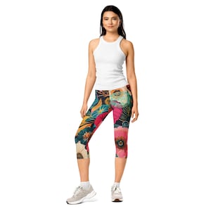 May include: A woman wearing a white tank top and floral print capri leggings. The leggings feature a colorful design with flowers in shades of pink, blue, and yellow against a dark background. The outfit is completed with white socks and gray sneakers.