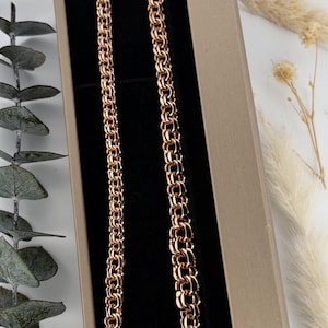 May include: A rose gold chain necklace featuring a dense, interlocking Byzantine link pattern. The necklace is displayed inside a rectangular, open-top box with a black velvet interior lining, showcasing the metallic luster of the links.